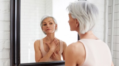 A mature woman with grey hair looking in the mirror while touching her neck and chest, which have stress hives