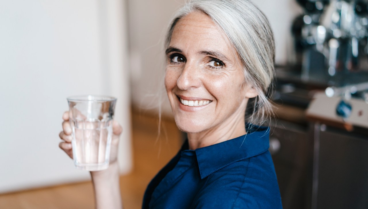 old woman with white hair tied back in a ponytail smiling with glass of water