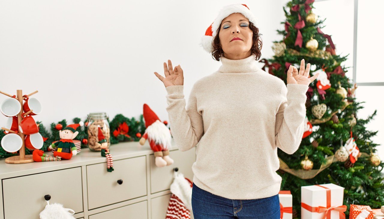 Middle aged hispanic woman standing by christmas tree with eyes closed doing meditation gesture with fingers.