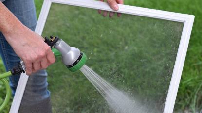 how to clean window screens: Closeup woman's hands washing window screens with water hose outside on green grass