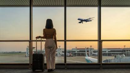 travel hacks for flying: Travel tourist standing with luggage watching sunset at airport window. Woman looking at lounge looking at airplanes while waiting at boarding gate before departure. Travel lifestyle. Transport and travel concept