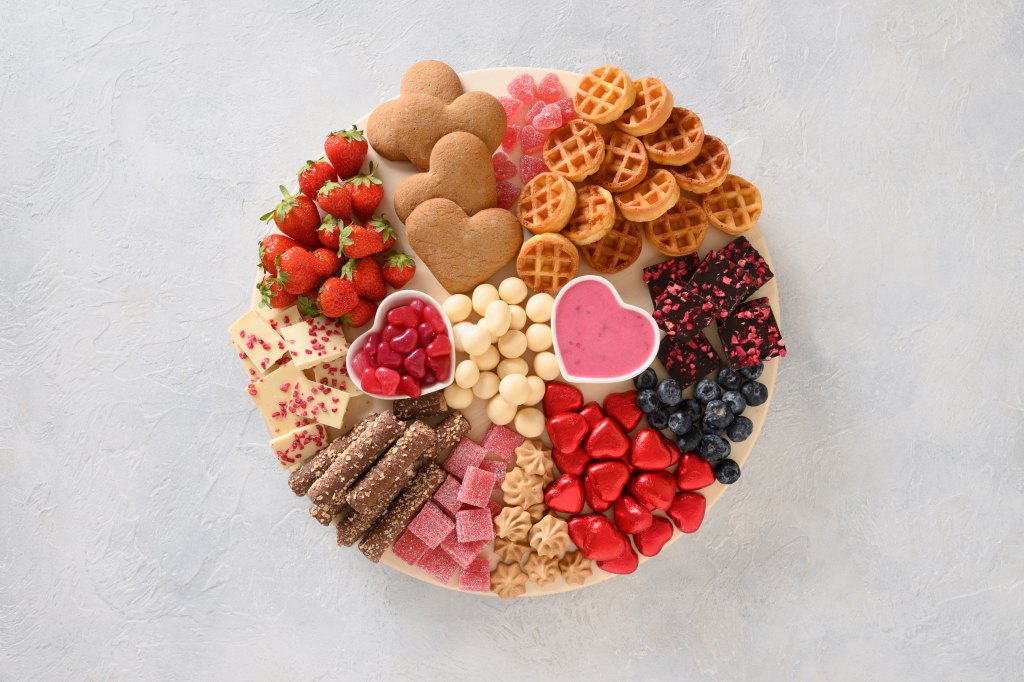 Charcuterie Valentines Day board with chocolate, red hearts, different sweets, strawberries, blueberries and candies on light gray background. View from above