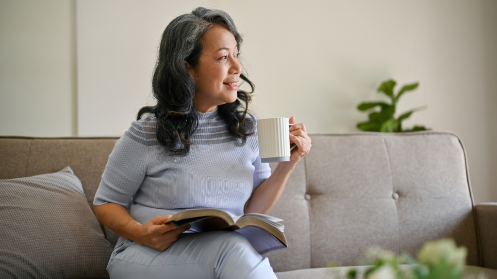 Woman smiling while sitting on a couch drinking coffee and holding a book, practicing dolce far niente