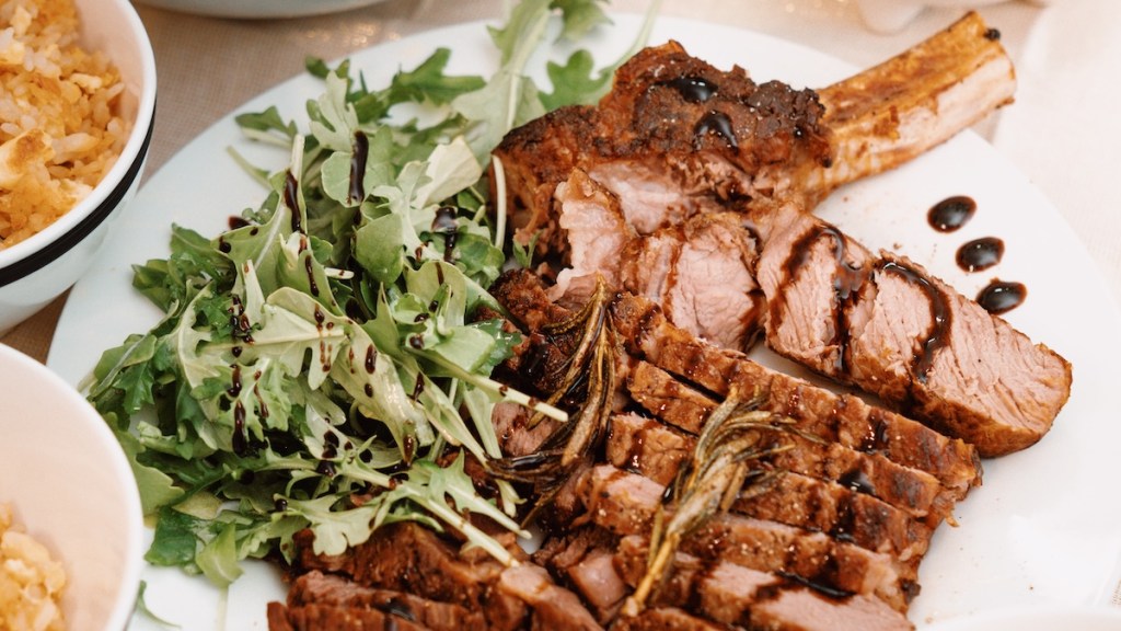 A plate features carnivore diet-friendly roast beef ribs on the bone cut in slices with some grilled rosemary leaves on the top. It also includes arugula salad.