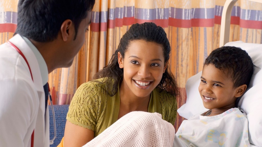 A child in a hospital bed next to their mother, who is speaking to a doctor