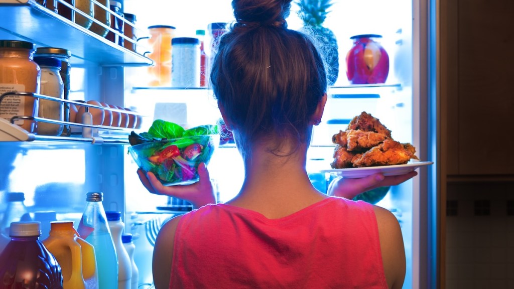 A young woman standing in front of the refrigerator, holding a bowl of fresh vegetable salad for a healthy diet on one hand and holding a plate of unhealthy fried chicken on the other. Making decision and choices for lifestyle and eating habit.