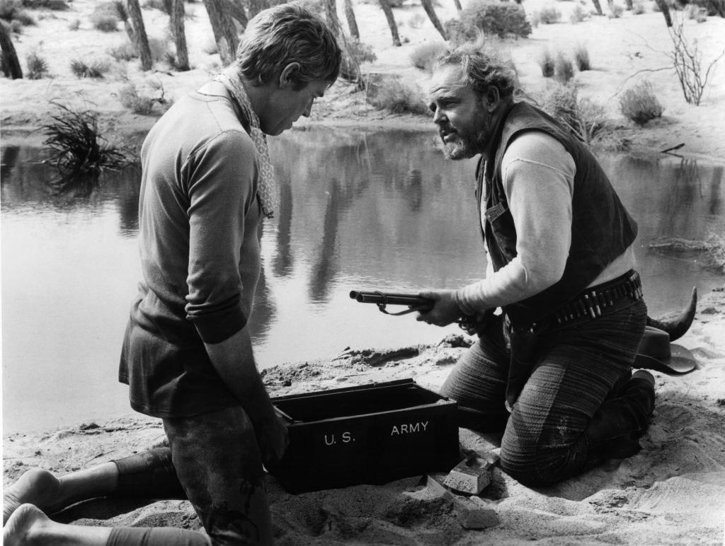 James Coburn has gun pulled on him by Carroll O'Connor in a scene from 'Waterhole #3', 1967. 