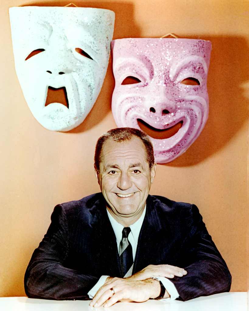 Jim Backus smiling as he poses beneath comedy and tragedy masks, which are mounted on the wall behind, circa 1960. 