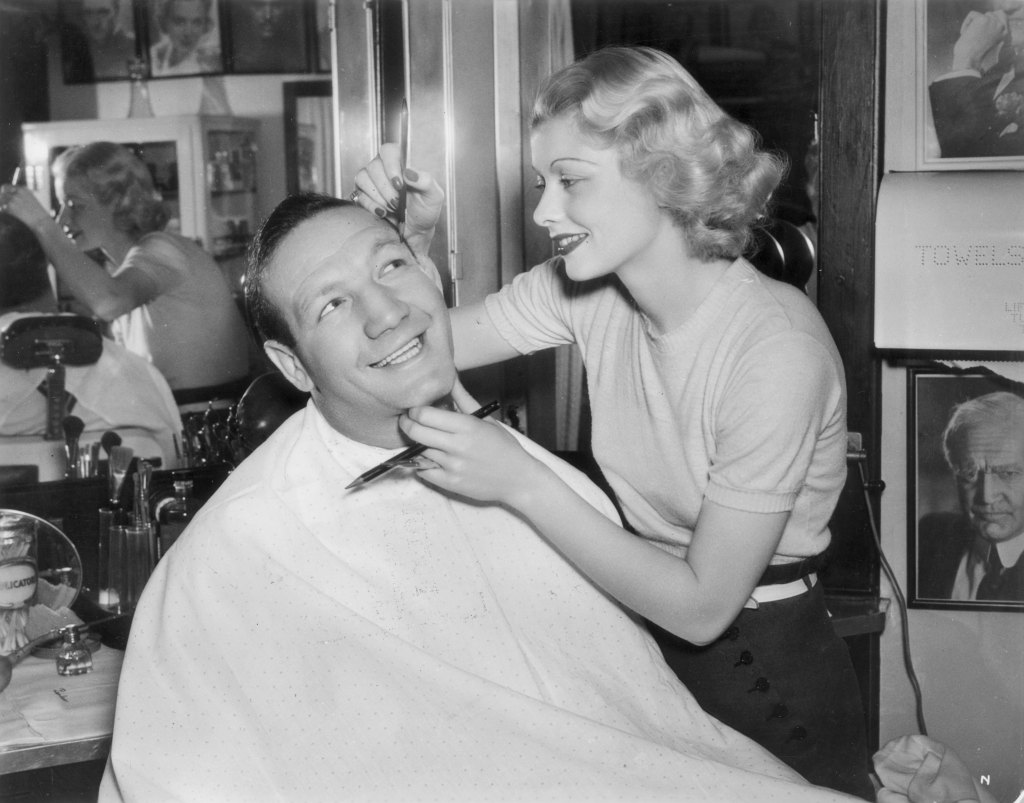 1936: American boxer Maxie Rosenbloom sits in a chair and smiles while American comedian Lucille Ball applies pencil to his eyebrow in a makeup room on the set of director Charles Vidor's film, 'Muss 'em Up'.