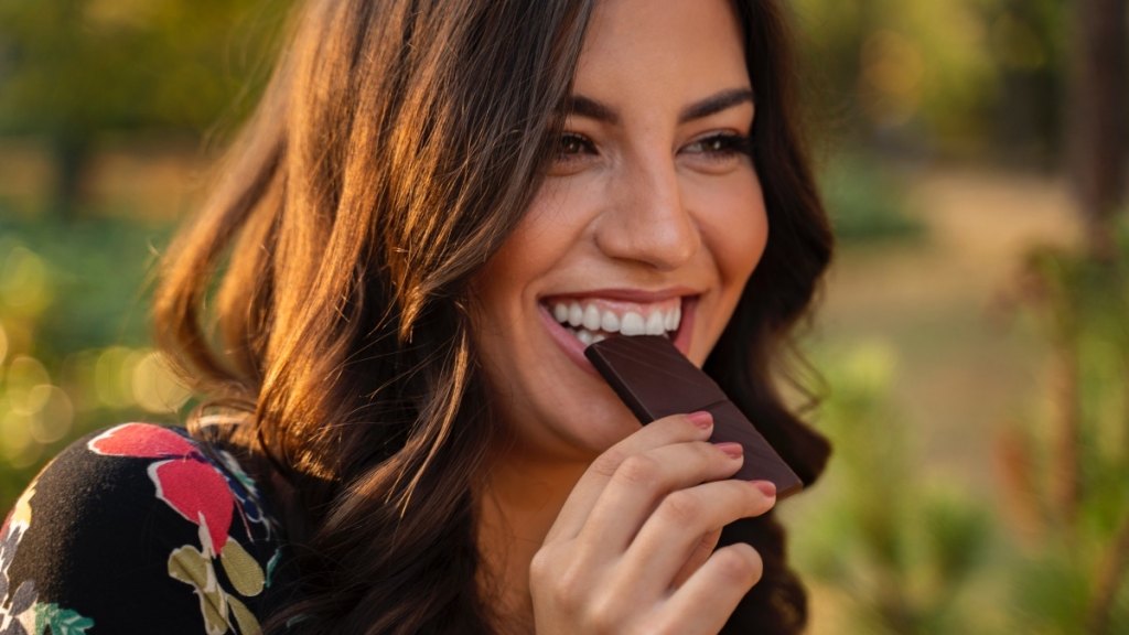 A happy woman eating a bark of dark chocolate to enjoy the health benefits