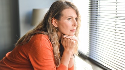A woman in an orange top looking at the window with her head in her hands while experiencing depression after an illness