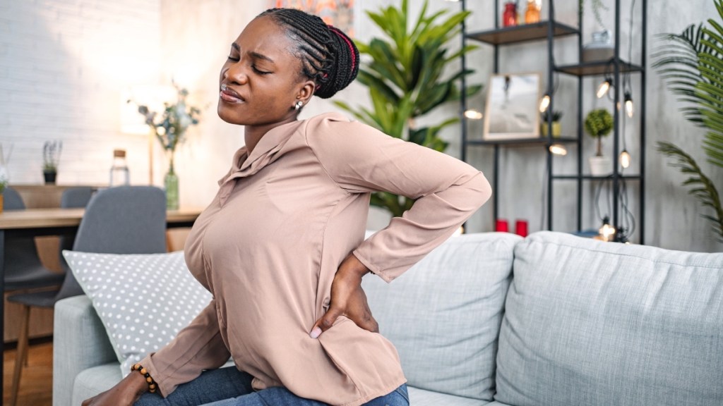 A woman sitting on a couch grimacing while pressing her hand onto her lower back where she has a pulled muscle