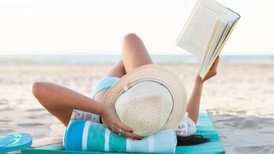 beast beach reads: Confident woman reads a book while sunbathing on a beach. She is wearing a straw hat. She is lying down on a blue beach towel with a rolled up towel underneath her head. The beach is in the distance.