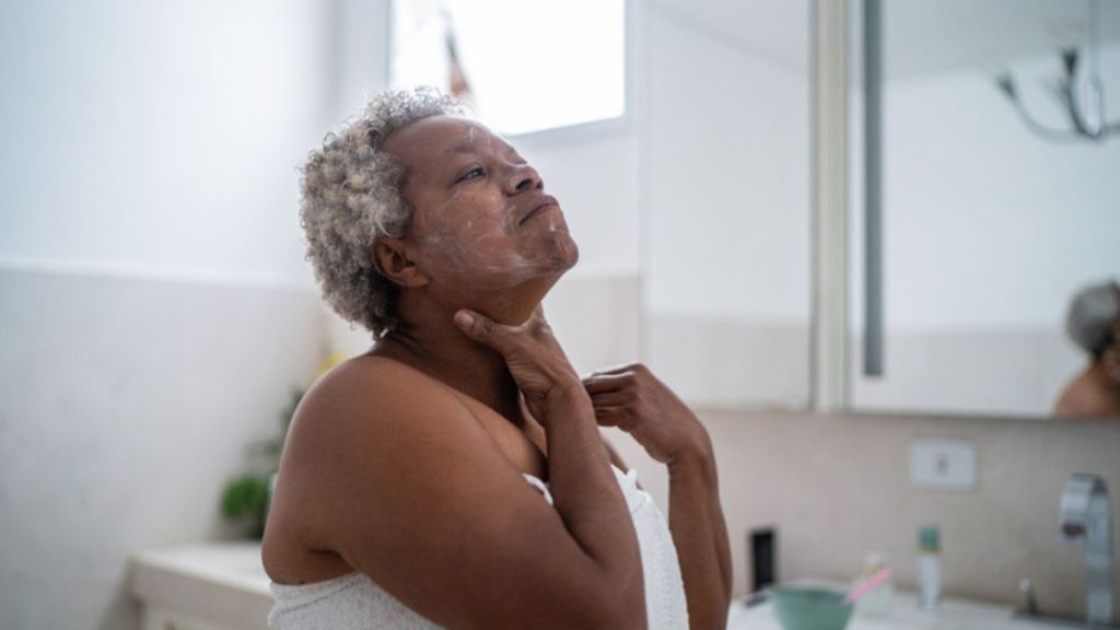 A mature Black woman applying moisturizer to her face while looking in the mirror