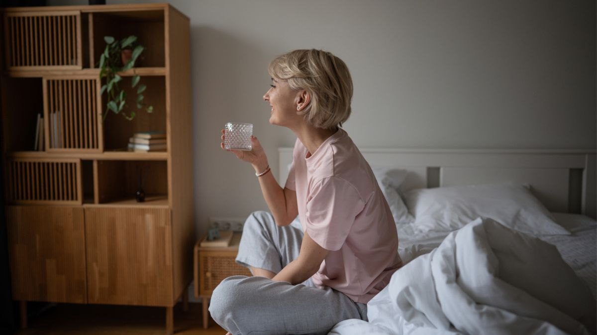 A woman sitting on a bed, drinking water as part of an analog wellness routine